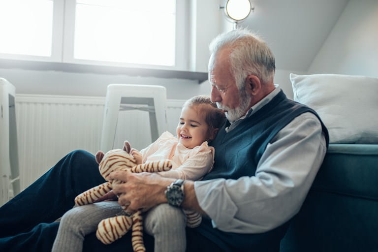 Grootvader die zijn kleinkind als begunstigde heeft aangeduid in een levensverzekeringscontract en met haar en haar knuffel speelt in de living.