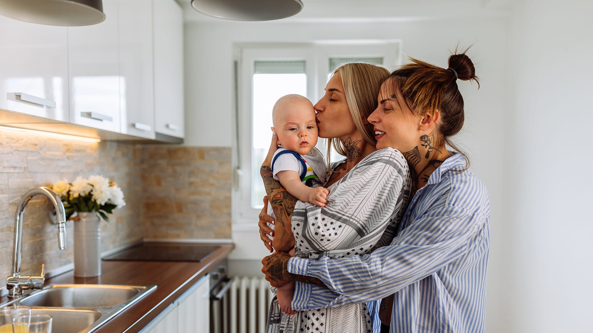 Deux femmes câlinent leur bébé dans la cuisine. Elles viennent de souscrire une assurance solde restant dû pour se protéger mutuellement en cas de décès.