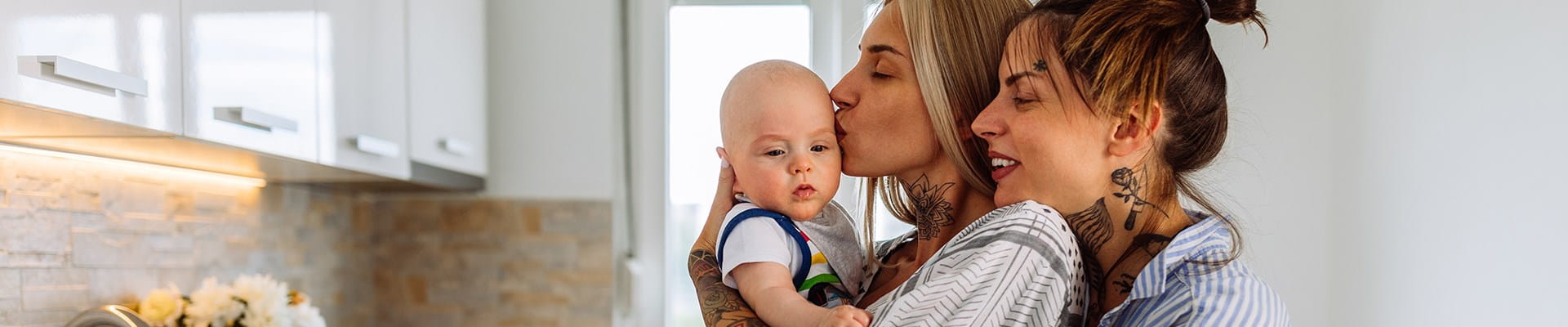 Deux femmes câlinent leur bébé dans la cuisine. Elles viennent de souscrire une assurance solde restant dû pour se protéger mutuellement en cas de décès.