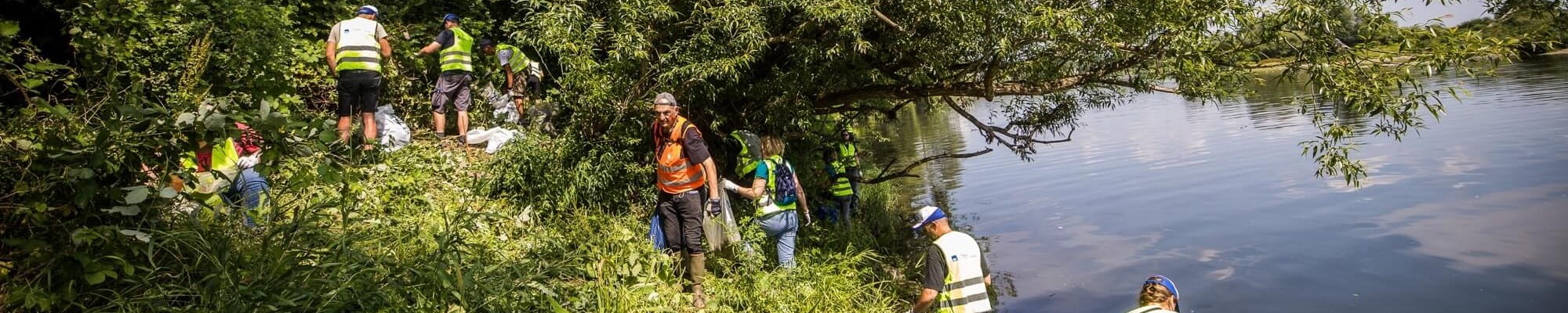 Vrijwilligers ruimen afval op langs een rivier na een overstroming, werkend in team in en rond het water met beschermende uitrusting.